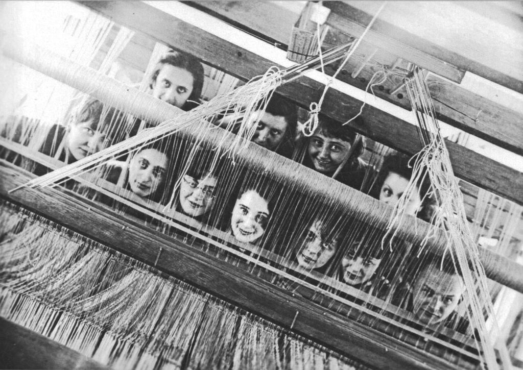 The ten Bauhaus weaving class participants and their teacher looking at the camera through a loom. 